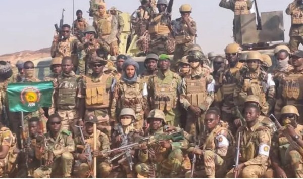 A large group of soldiers in camouflage uniforms with helmets and gear pose together outdoors, some holding weapons and a green flag.
